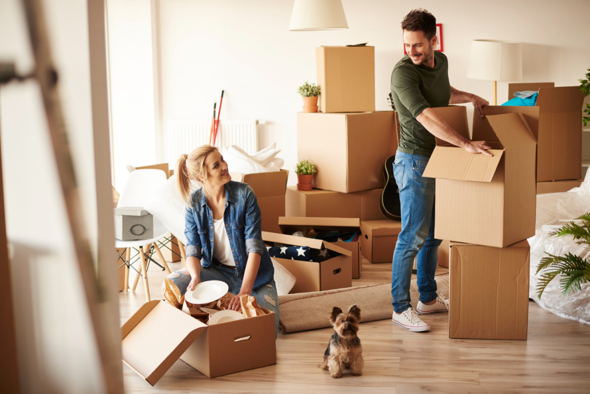 A couple unpacking cardboard boxes in their new home, with their dog sitting on the floor